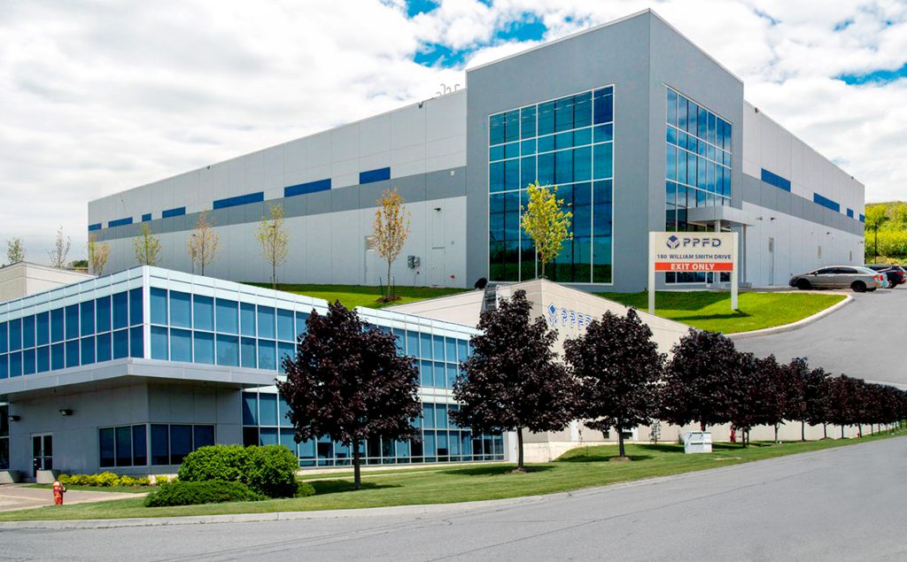 The modern headquarters of PPFD, a large building with extensive glass windows, located at 150 William Smith Drive. The building is surrounded by neatly maintained landscaping, including rows of trees and shrubs. The clear signage in front indicates the company name and address.