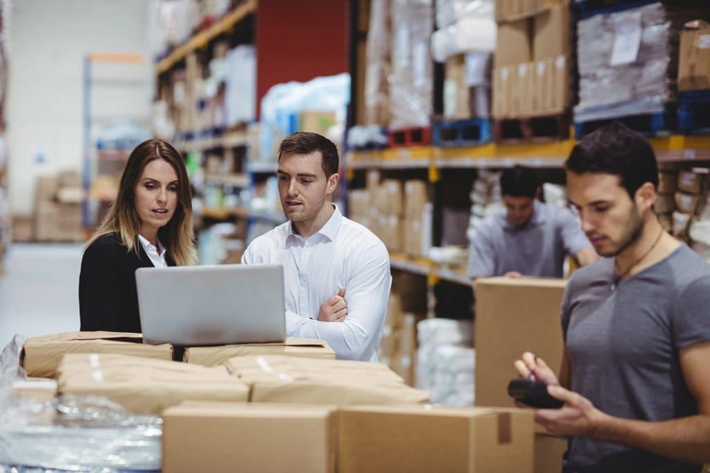 People working in a warehouse; two are focused on a laptop amid stacked boxes, while another person uses a handheld device in the foreground.