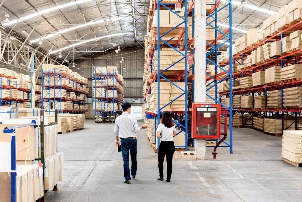 Two people walking through a large warehouse with tall shelves filled with stacked boxes.