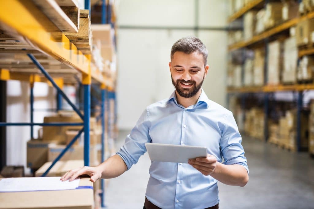 A man with a beard and wearing a light blue shirt stands smiling in a warehouse, holding a tablet. He is surrounded by shelves filled with boxes, indicating an organizational or inventory setting.