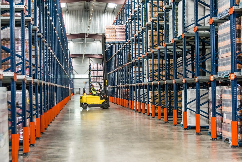 A yellow forklift moves through a large warehouse aisle lined with tall blue and orange racks filled with stacked pallets. The warehouse has a high ceiling and is well-lit, with a long perspective down the aisle.