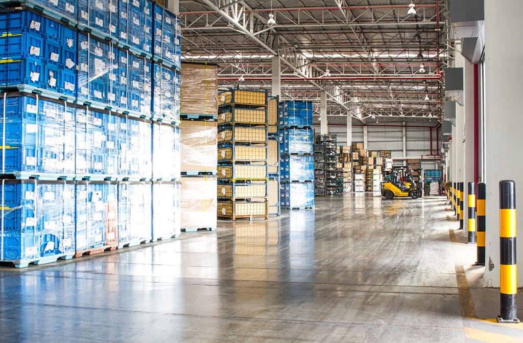 A spacious warehouse with tall stacks of blue and brown pallets on the left. A forklift is seen moving boxes on the right. The floor is shiny and clean, with yellow and black safety barriers along the walls.